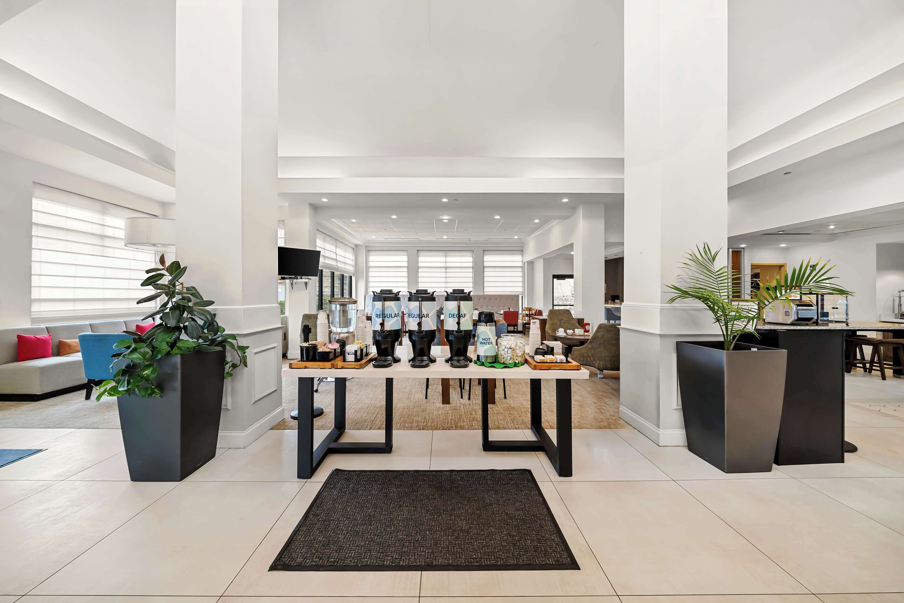 Eine großzügige Lobby mit einem zentralen Tisch, auf dem verschiedene Kaffeemaschinen präsentiert werden, einer großen Topfpflanze und einer modernen Sitzecke mit Sofa und Sesseln.