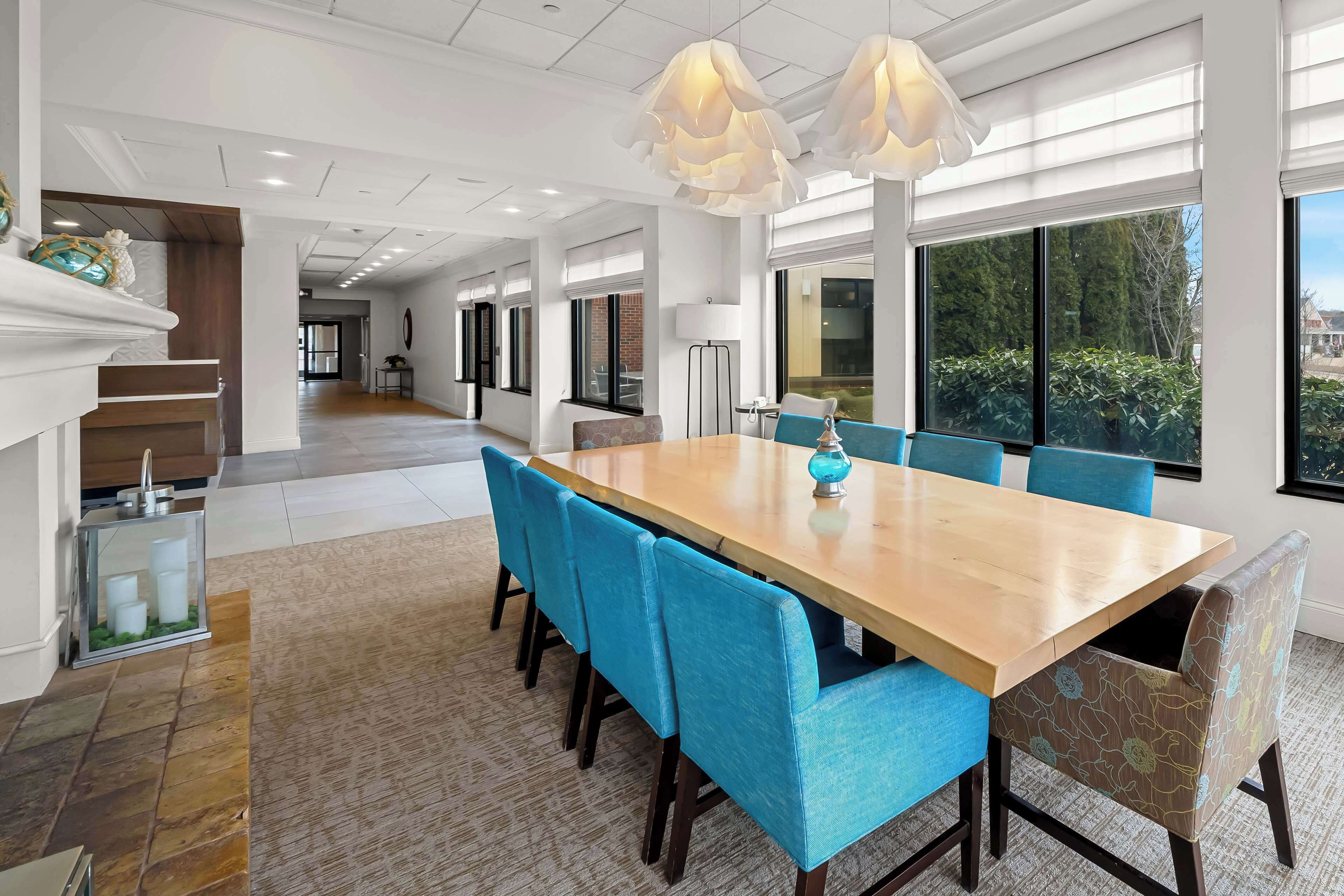 Un vaste espace dédié à la salle à manger, avec une longue table en bois, des chaises recouvertes d’un tissu bleu et une grande fenêtre donnant sur l’extérieur.