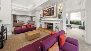 A spacious living room with a fireplace, a large painting, and a modern kitchen in the background. - Hilton Garden Inn Plymouth (Plymouth)