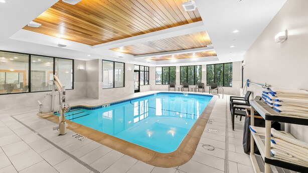 An indoor swimming pool with wooden ceiling panels, clear glass walls, and a seating area with tables and chairs. - Hilton Garden Inn Plymouth (Plymouth)