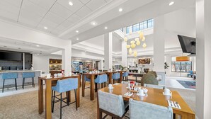 A modern hotel lobby with multiple dining tables, chairs, and a kitchen area in the background. - Hilton Garden Inn Plymouth (Plymouth)