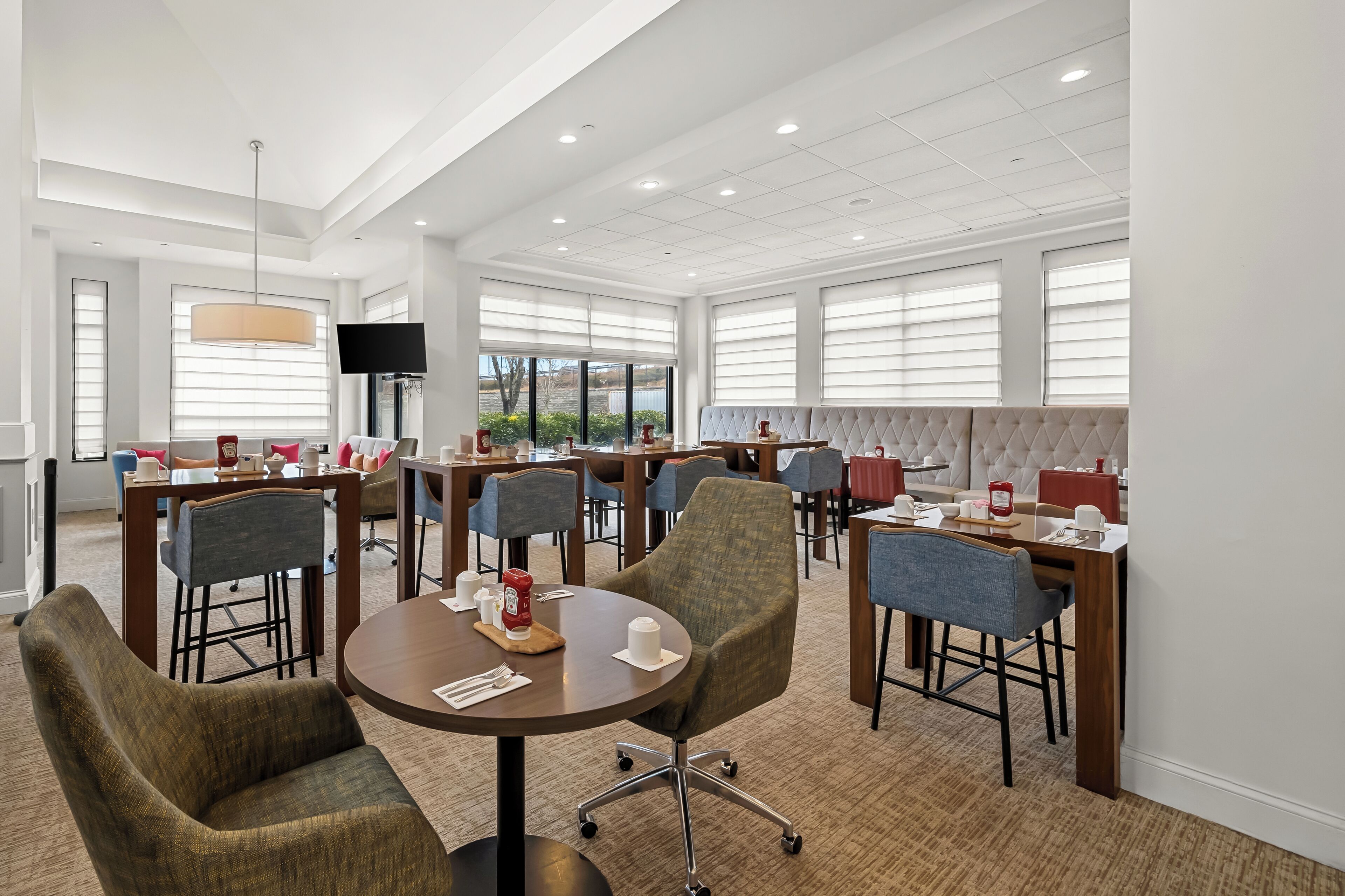 Ruime hotel eetruimte met meerdere tafels en stoelen, een aan de muur bevestigde televisie en grote ramen die voor natuurlijk licht zorgen.