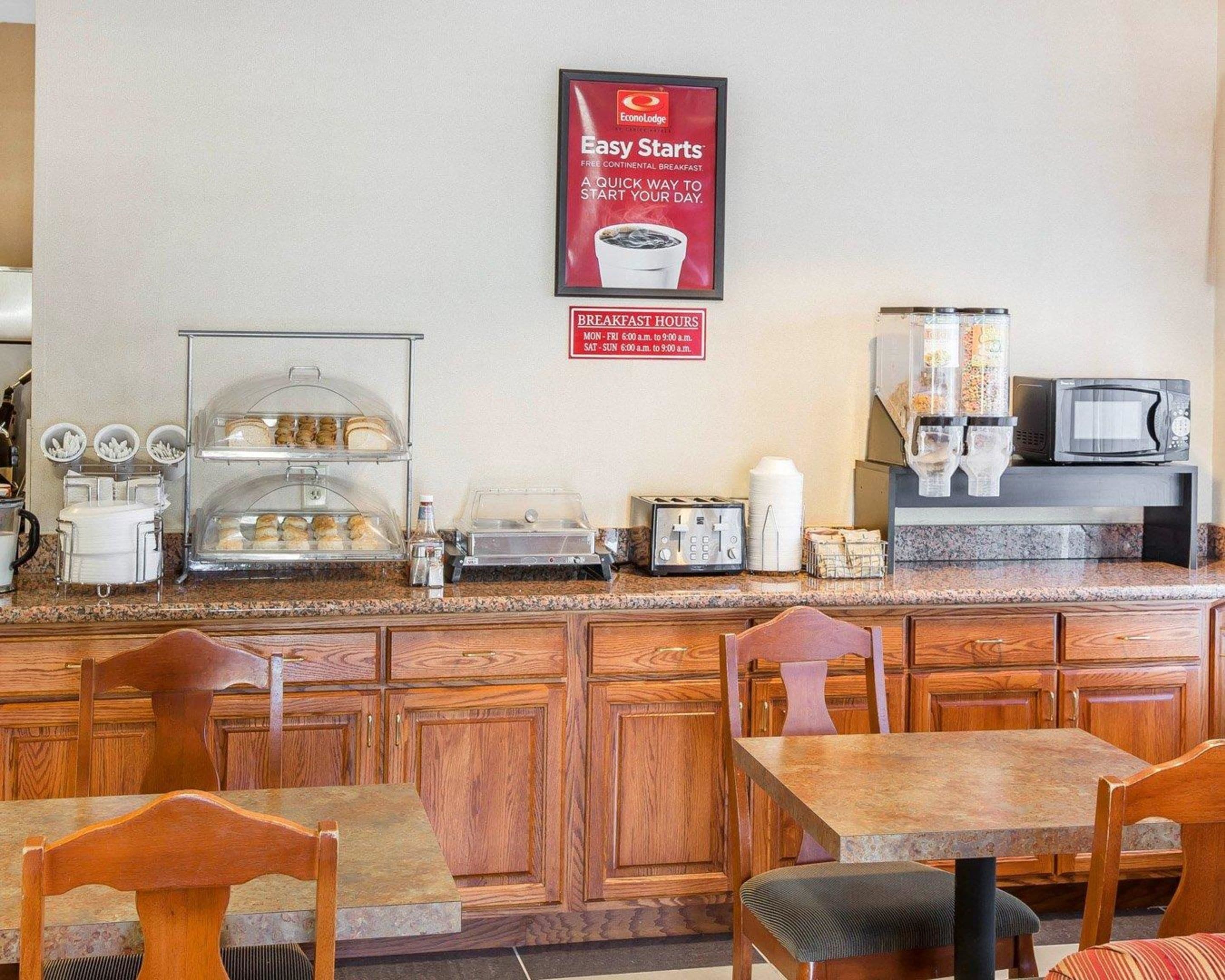 Un área de desayuno con un cartel que dice 'EconoLodge Easy Starts' y un cartel de 'Horario del Desayuno'. Hay dos mesas y sillas, una cafetera, un microondas y una barra con alimentos.