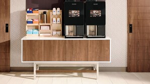 A coffee station with two coffee machines, a shelf with coffee cups, and a storage area with boxes and a kettle. - Holiday Inn Express & Suites South - Lincoln by IHG (Lincoln)
