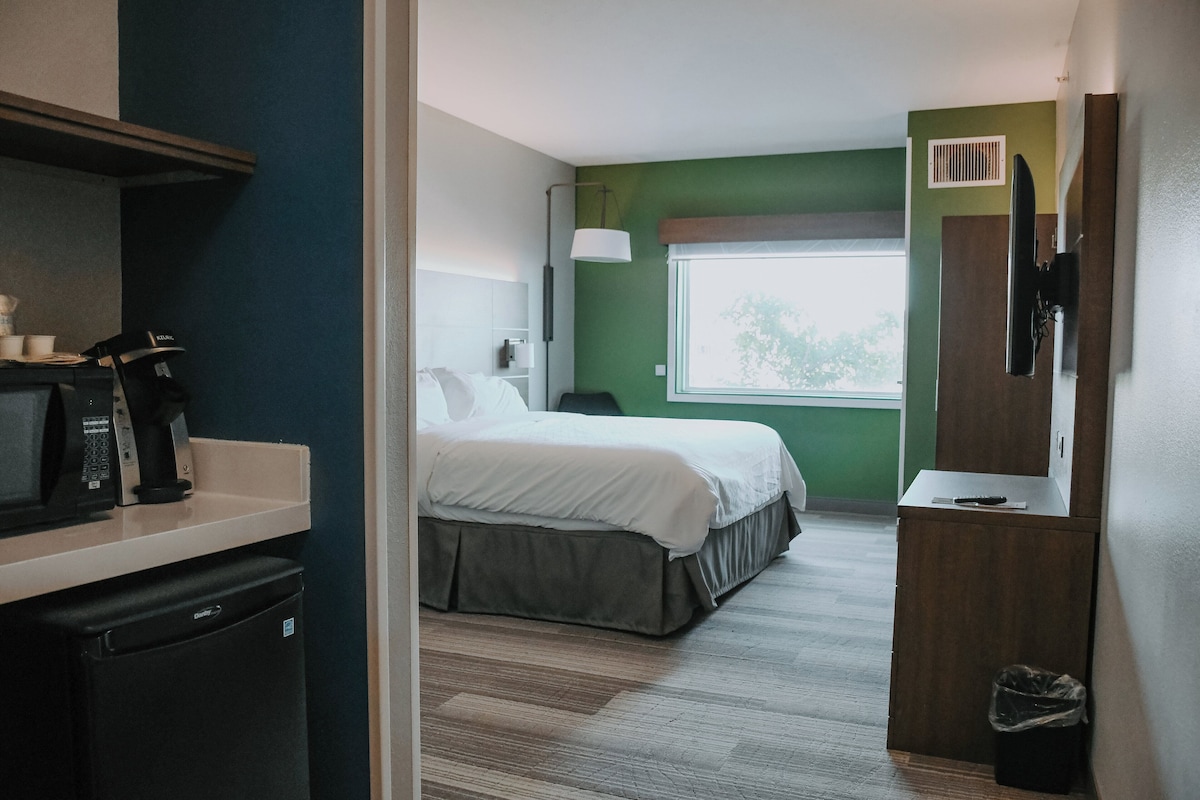 A hotel room with a bed, a desk with a microwave, a trash bin, and a window with a view of trees.