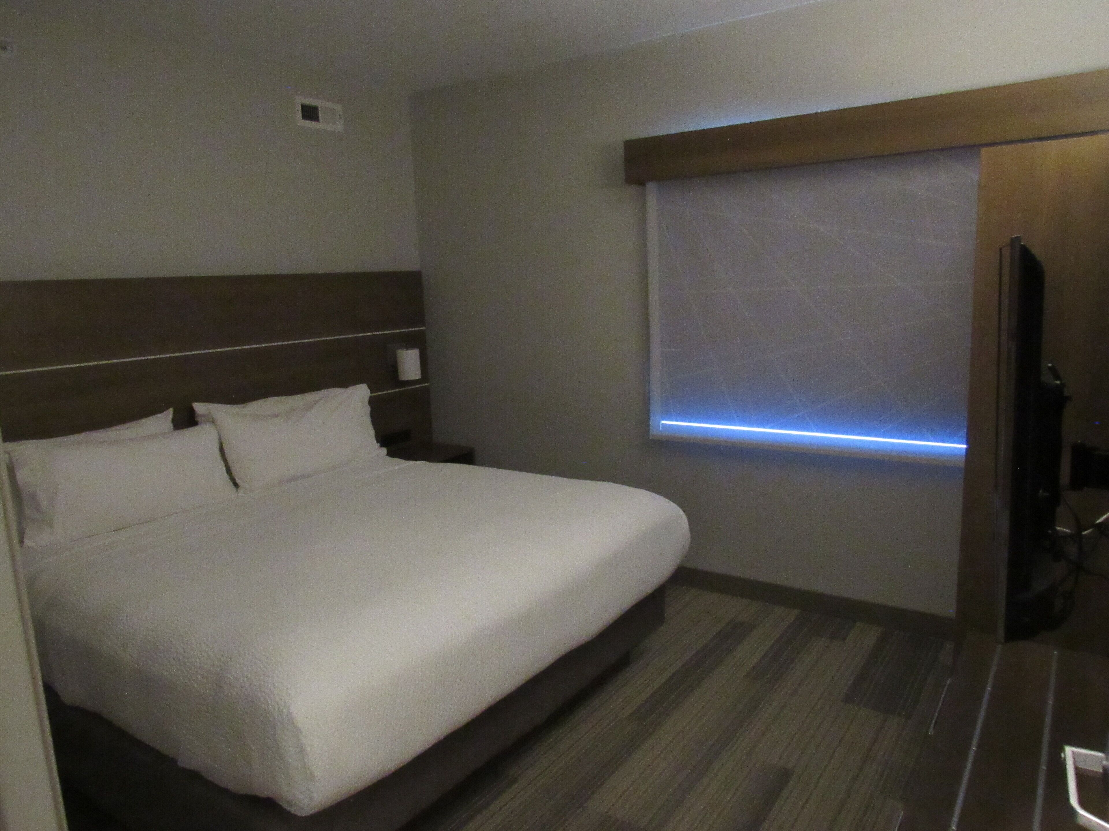 Une chambre d’hôtel avec un grand lit, une tête de lit en bois, un téléviseur fixé au mur et une fenêtre laissant filtrer une lumière bleue.