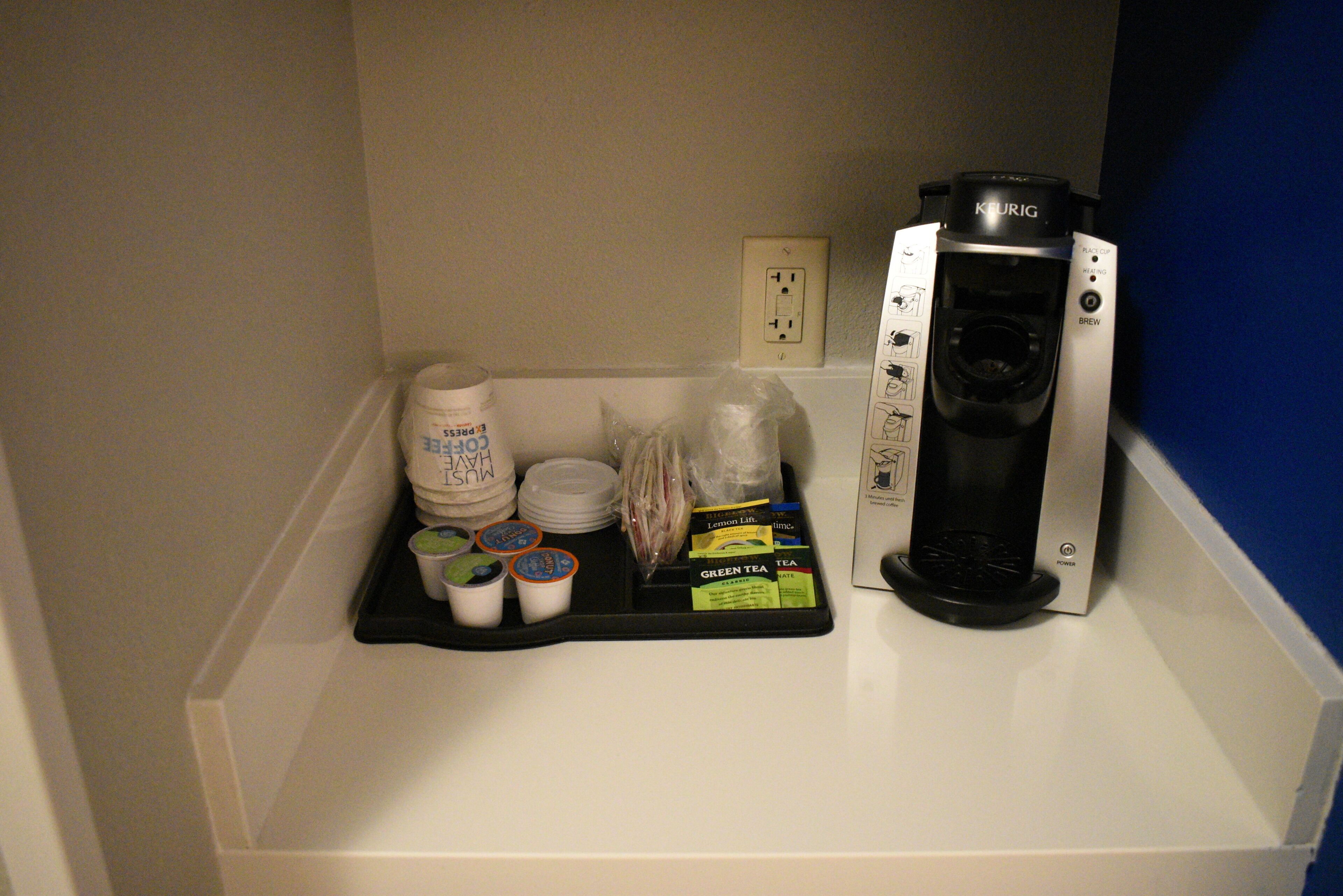 Une machine à café est posée sur une étagère blanche, à côté d’un plateau contenant des dosettes de café, des tasses et un paquet de sucre.