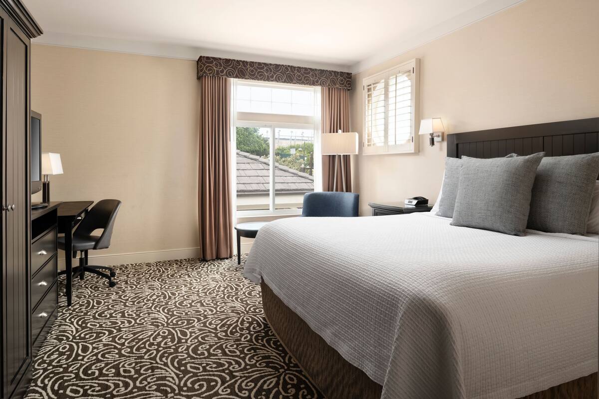 A hotel room with a large bed, a desk with a chair, a desk lamp, a nightstand, and a window with curtains.