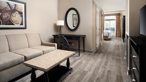 A hotel room with a sofa, a small table, a desk, a chair, a TV, a mirror, and a wardrobe. - West Inn & Suites (Carlsbad)