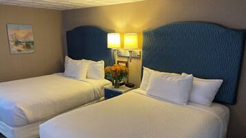A hotel room with two beds, each with white bedding and blue headboards. There is a nightstand between the beds with a lamp and a vase of flowers.
