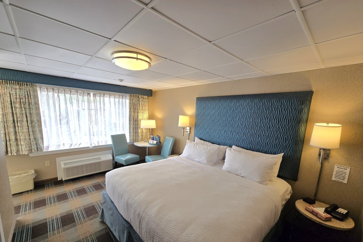 A hotel room with a large bed, two armchairs, a small table with a lamp, and a window with curtains.