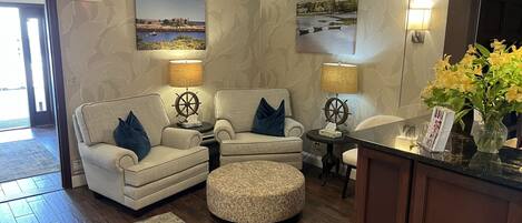A cozy living room with a sofa, armchair, ottoman, and a small table with a ship wheel lamp. There is a wall-mounted picture, a vase with flowers, and an exit sign.