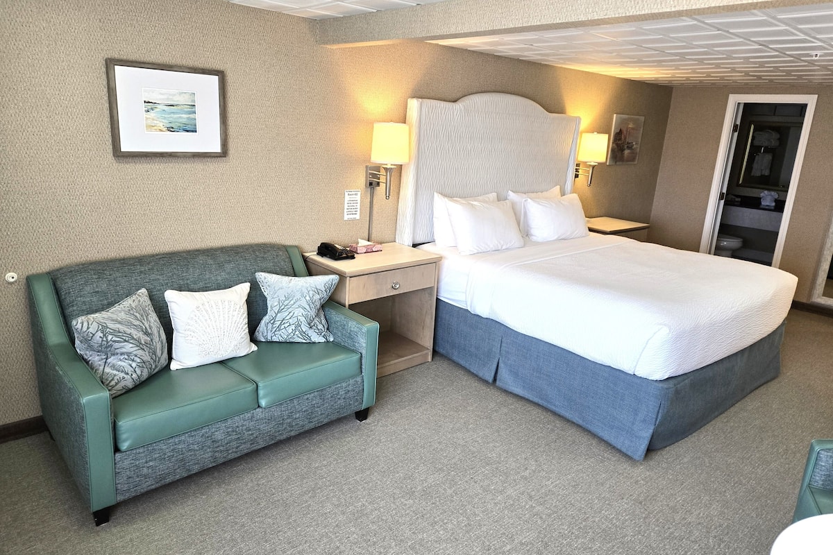 A hotel room with a bed, a green sofa with patterned cushions, a nightstand with a lamp, and a framed picture on the wall.