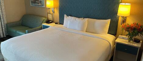 Una habitación de hotel con una cama grande, una mesita de noche con lámpara, una pequeña mesa con un florero y un sillón verde.