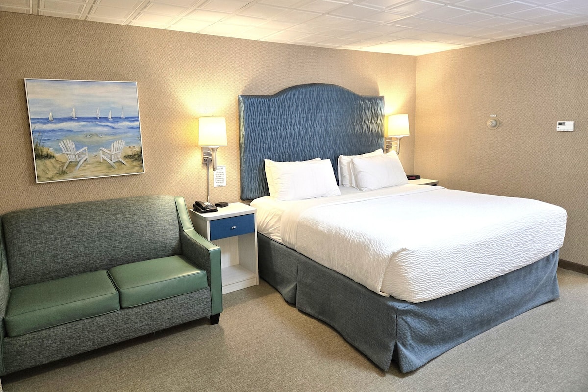 A hotel room with a bed, a nightstand, a lamp, a painting of a beach scene, and a green sofa.