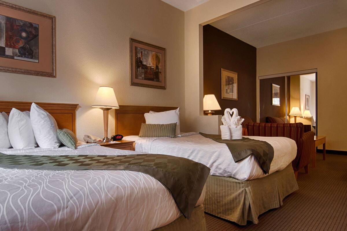 A hotel room with two beds, a nightstand with a lamp, a phone, and a wall clock. There are framed pictures on the wall and a chair in the corner.