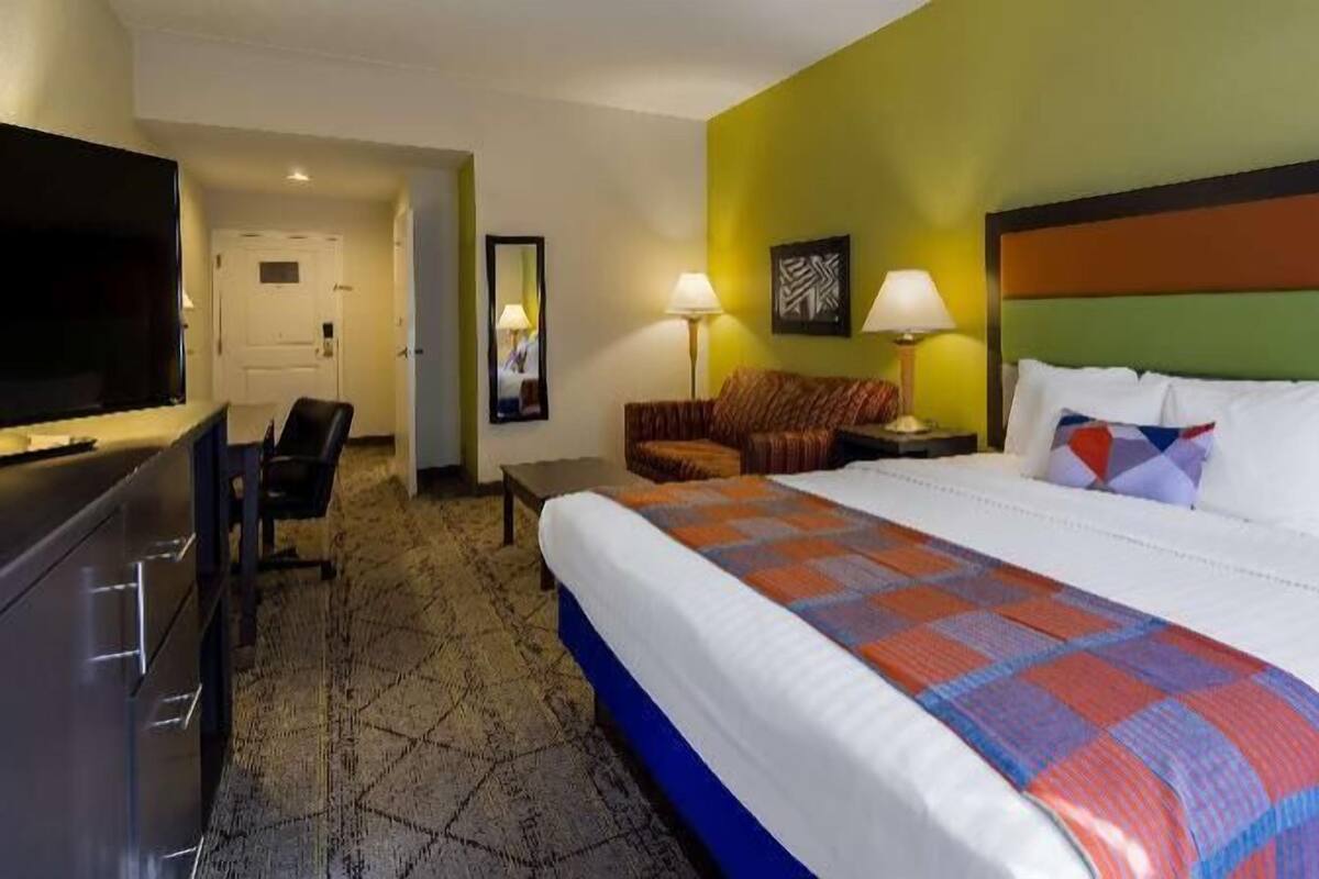A hotel room with a bed, a desk with a TV, a chair, a lamp, and a door.