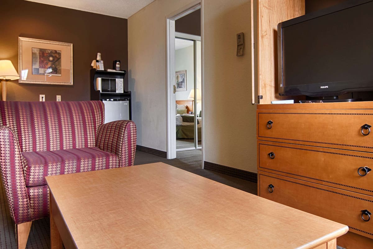 A hotel room with a wooden table, a patterned armchair, a television on a wooden cabinet, and a kitchenette with a microwave.