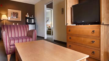 Une chambre d’hôtel avec une table en bois, un fauteuil à motifs, un téléviseur posé sur un meuble en bois et une kitchenette équipée d’un four à micro-ondes.