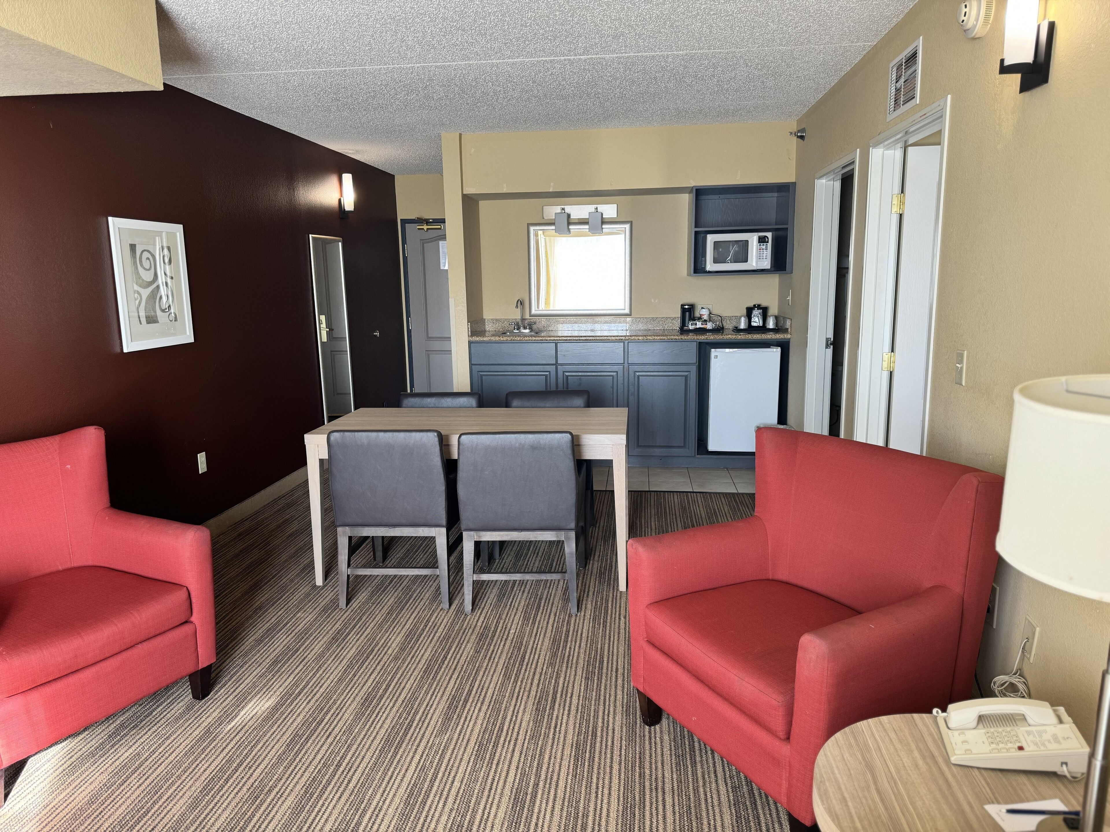 A compact hotel room with a dining area, kitchenette, and two red armchairs.