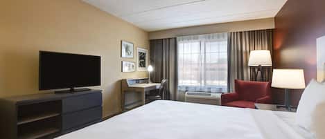 A hotel room with a bed, a desk, a television, two chairs, a window with curtains, and a lamp.