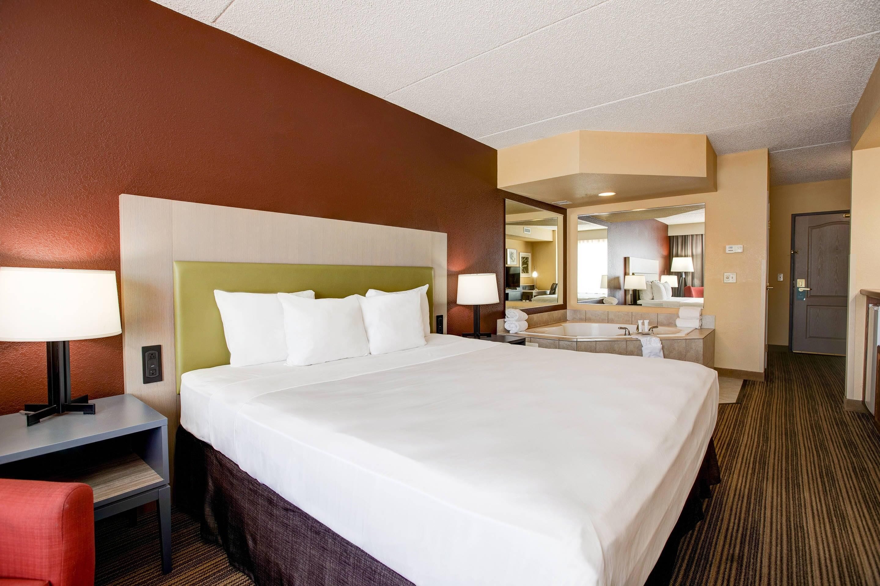 Ein Hotelzimmer mit einem großen Bett, Nachttischlampen, einem Schreibtisch, einem Sessel, einem Badezimmer mit Waschbecken und Spiegel sowie einer Tür.