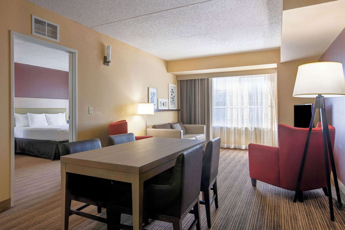 A hotel room with a bed, a dining table with chairs, a red armchair, a lamp, and a large window with curtains.