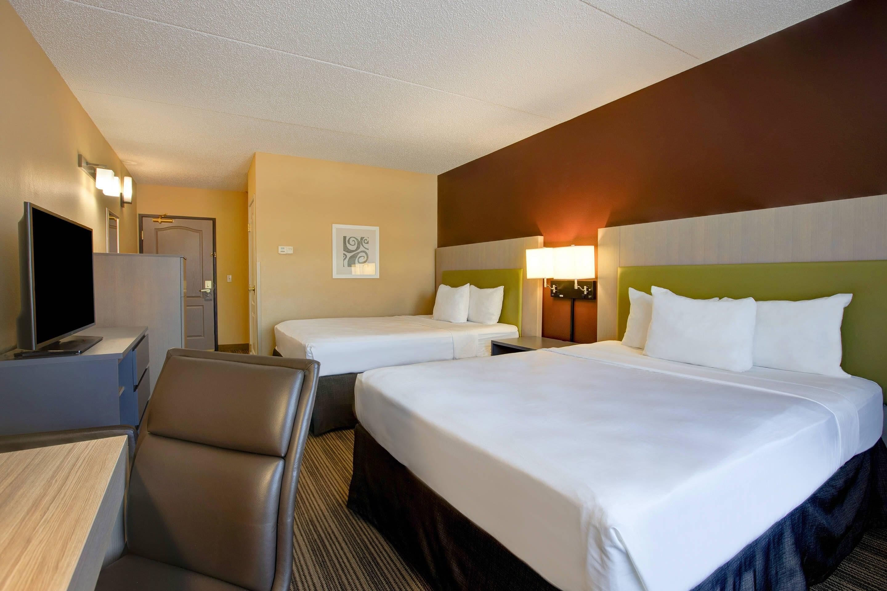 A hotel room with a large bed, two smaller beds, a desk with a flat-screen TV, a chair, and a wall-mounted lamp.