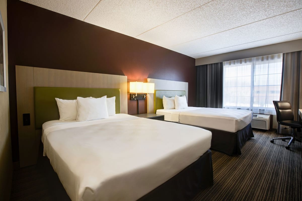 A hotel room with two beds, a desk, a chair, a lamp, and a window with curtains.