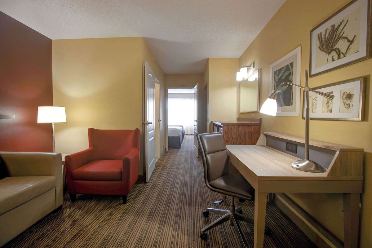 A hotel room with a desk, two chairs, a lamp, and framed artwork on the wall.