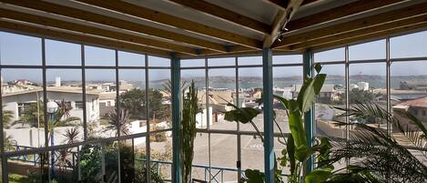 Un espace extérieur abrité offrant une vue sur le paysage urbain, avec un plafond en bois, de grandes fenêtres et un coin salon comprenant une table et des chaises.