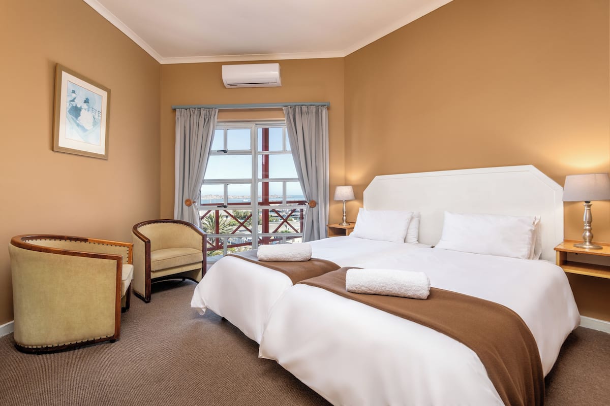 A hotel room with a large bed, two armchairs, a window with curtains, a framed picture, and a lamp on the nightstand.