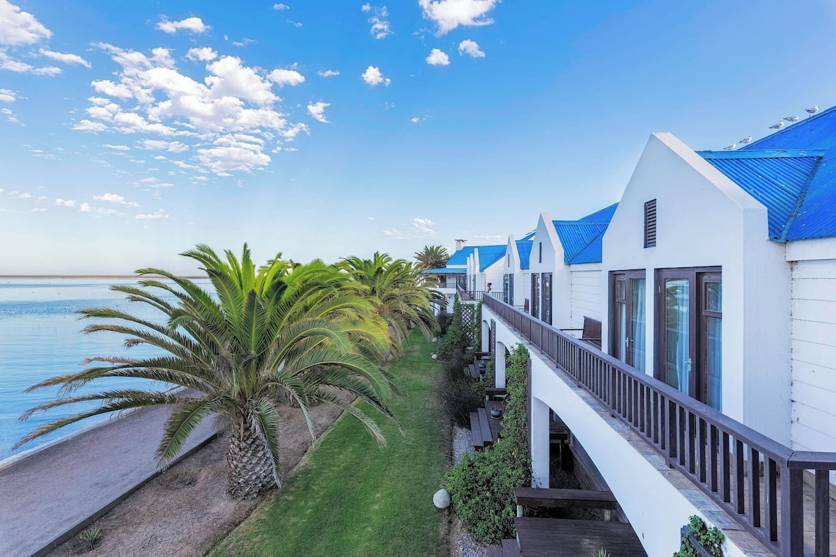 A coastal hotel with white buildings and blue roofs, a grassy area with palm trees, and a view of the sea.