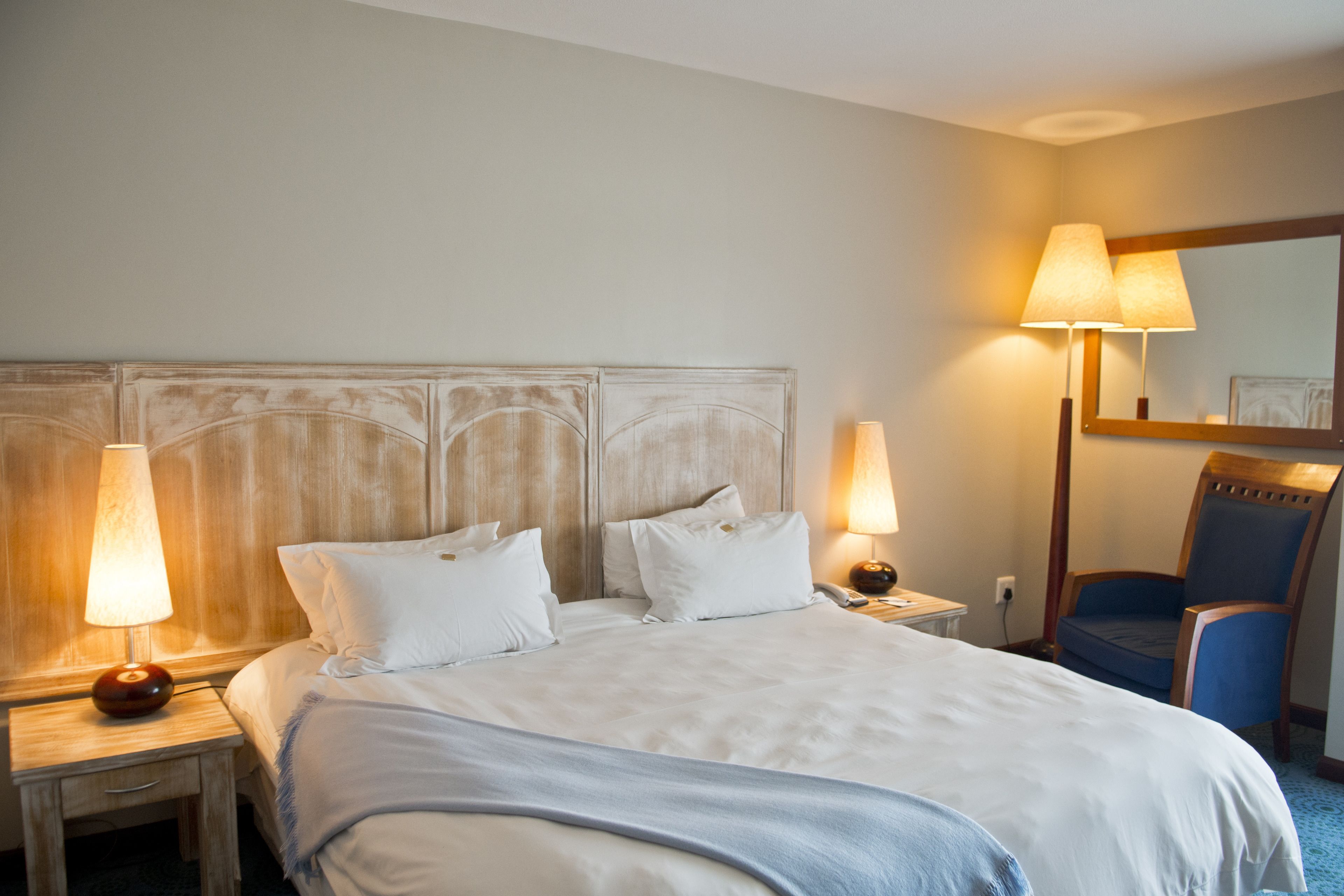 A hotel room with a large bed, two bedside lamps, a wooden headboard, a chair, and a mirror on the wall.