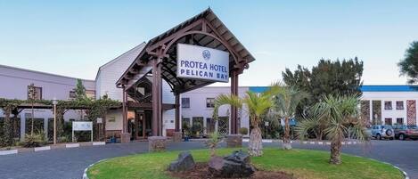 The entrance of Protea Hotel Pelican Bay with a prominent sign, a landscaped garden with rocks and palm trees, and a paved driveway.