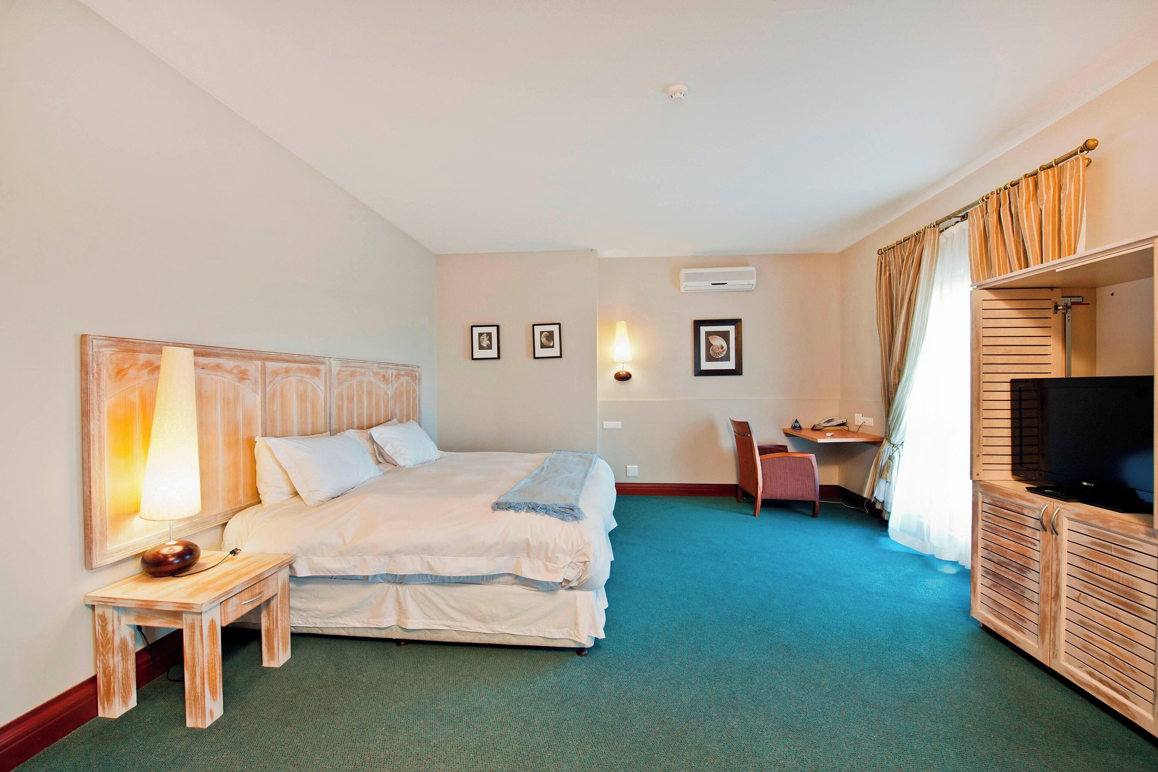 Quarto de hotel com cama de casal, escrivaninha com cadeira, televisão e mesinha de cabeceira com abajur.