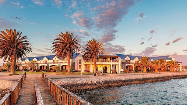 A resort with a wooden boardwalk leading to a building with multiple balconies, palm trees, and a body of water. - Protea Hotel by Marriott Walvis Bay Pelican Bay (Walvis Bay)