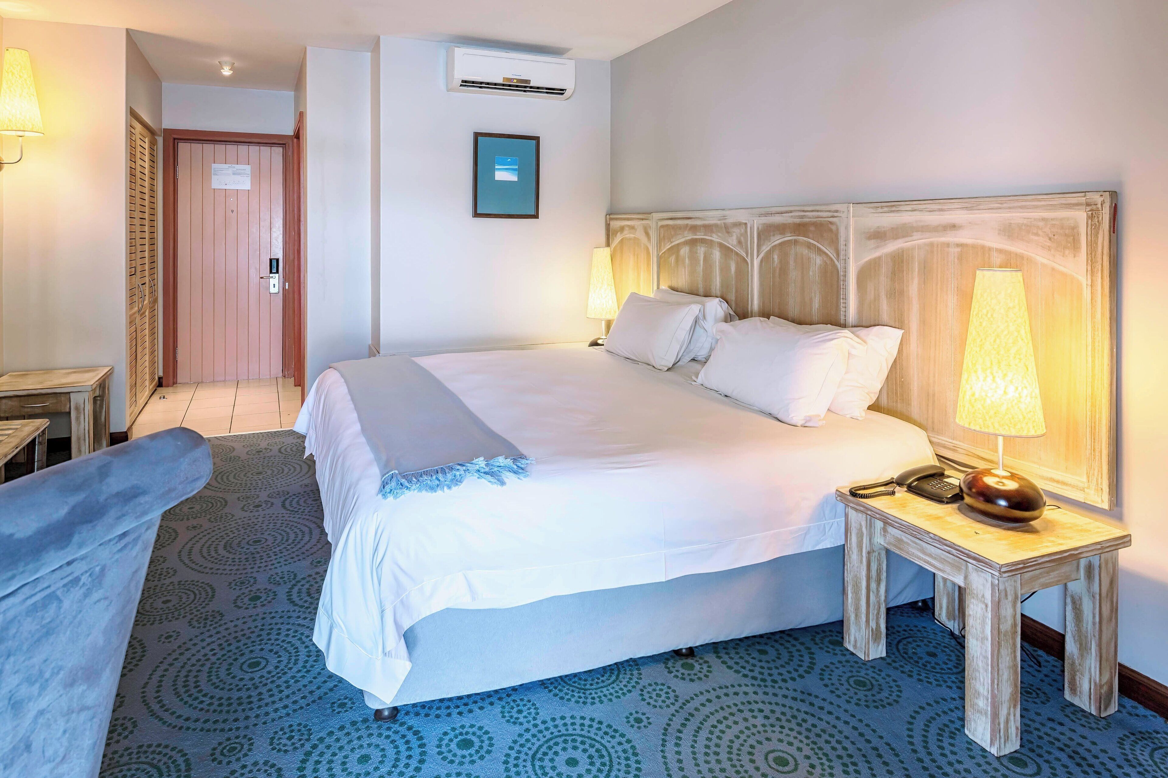 A hotel room with a large bed, a bedside table with a lamp, a patterned carpet, and a door with a sign.