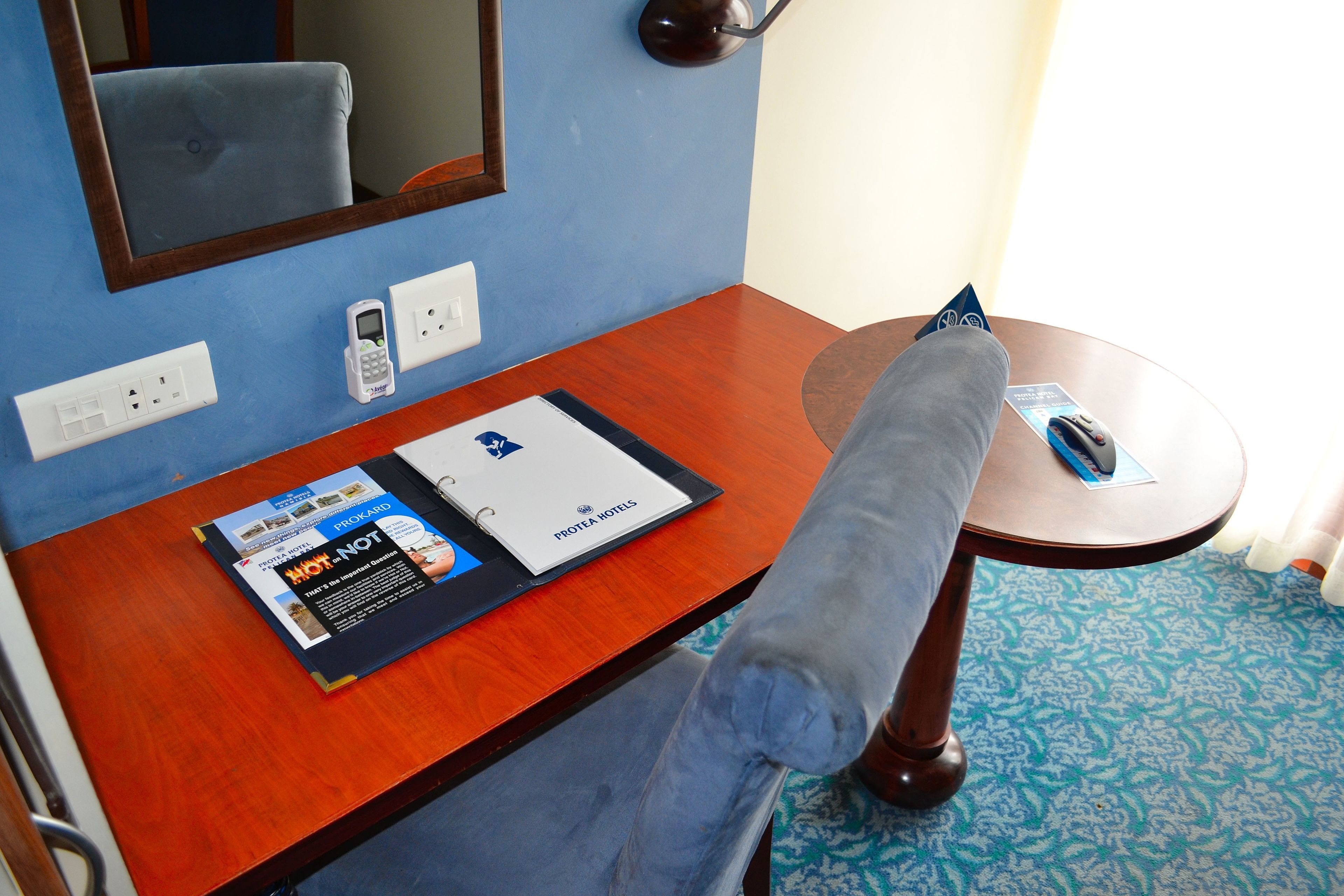 Habitación de hotel con un escritorio de madera, una silla, un espejo, un teléfono y un folleto sobre el escritorio.