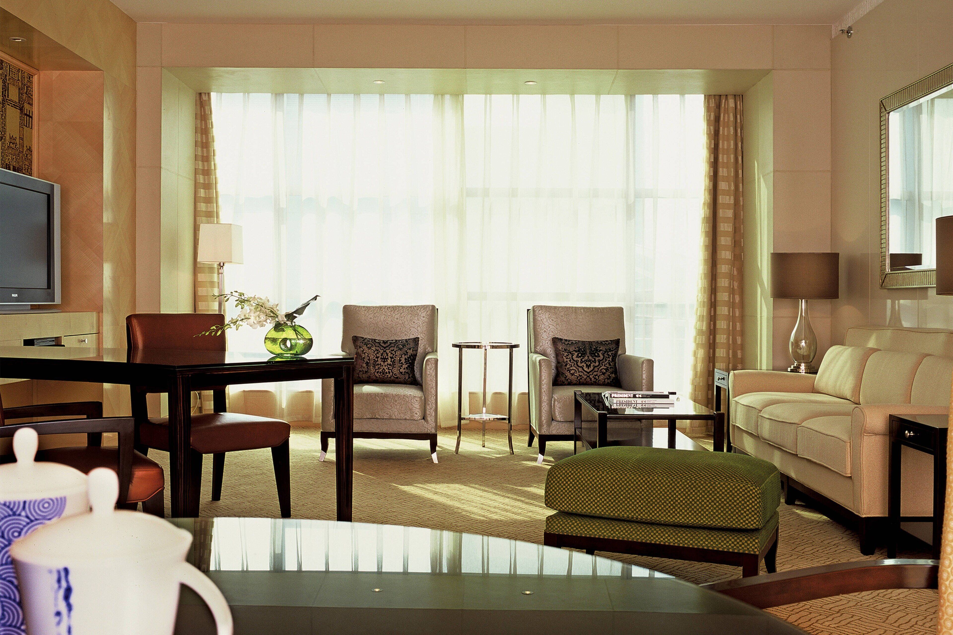 A hotel room with a television, two armchairs, a sofa, a dining table, and a green ottoman.