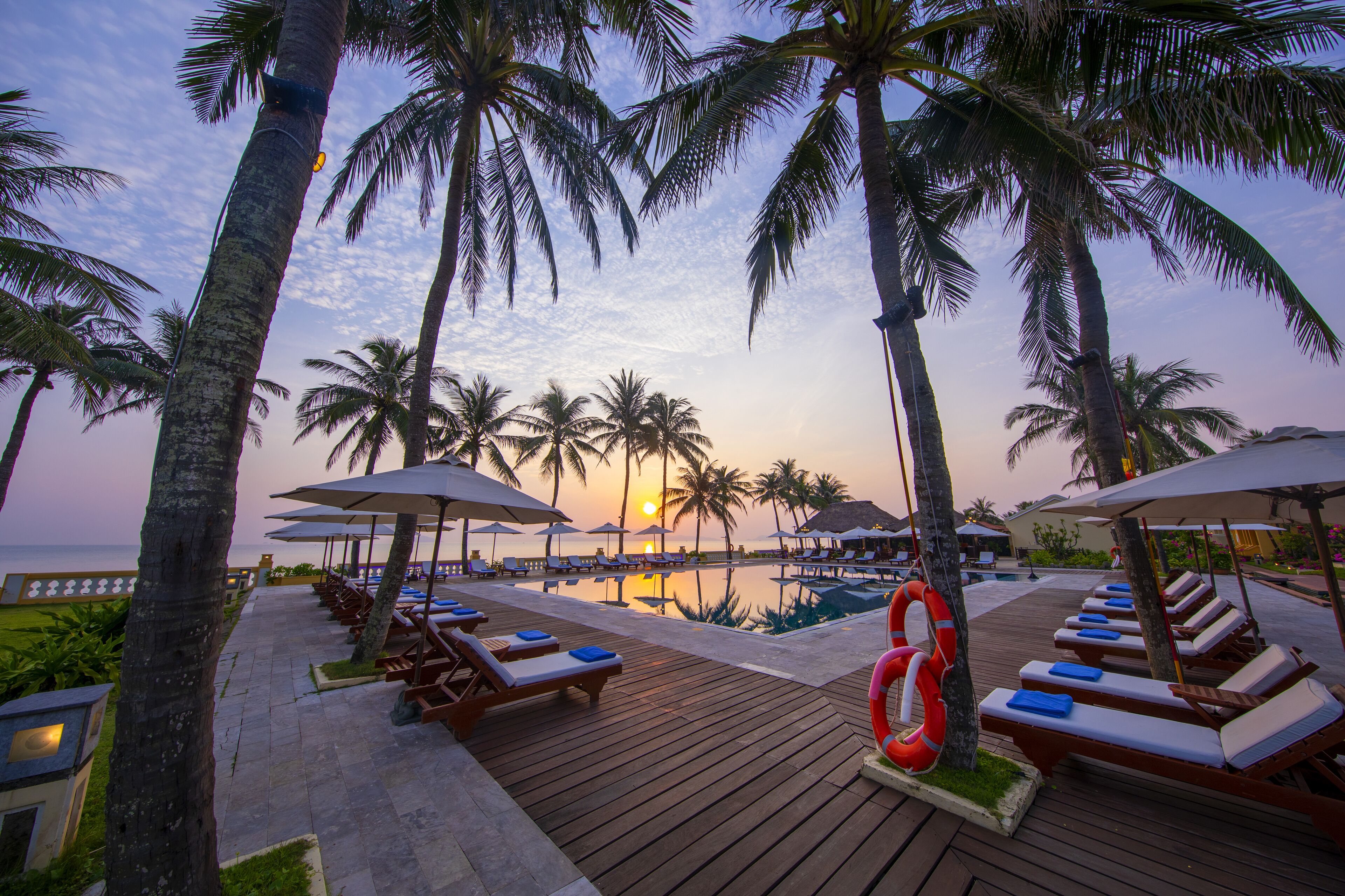 Terraza de madera con sillones y sombrillas, con vista a una piscina y palmeras al atardecer.