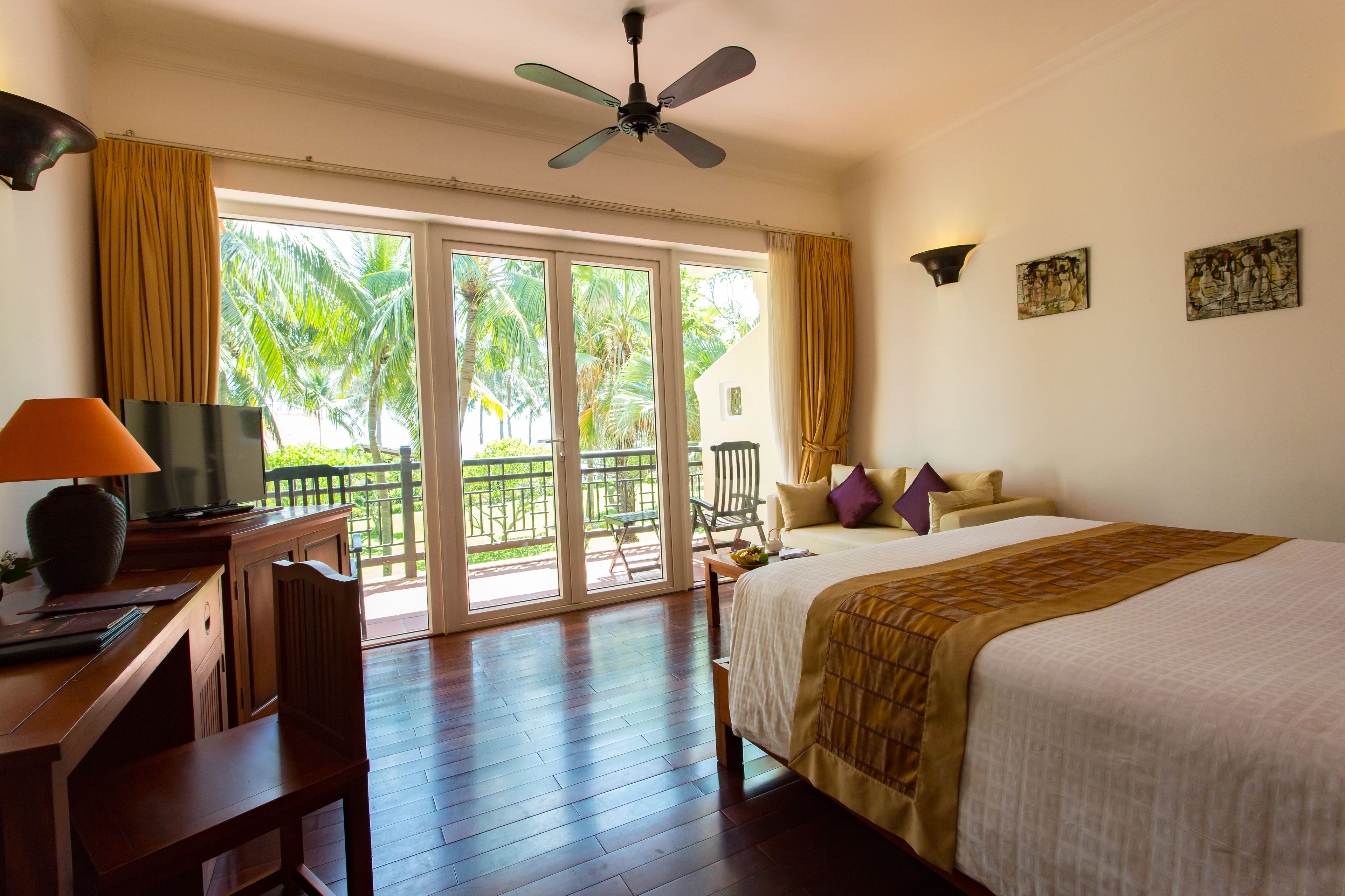 A hotel room with a bed, desk, and chair, a ceiling fan, and a view of palm trees through the window.