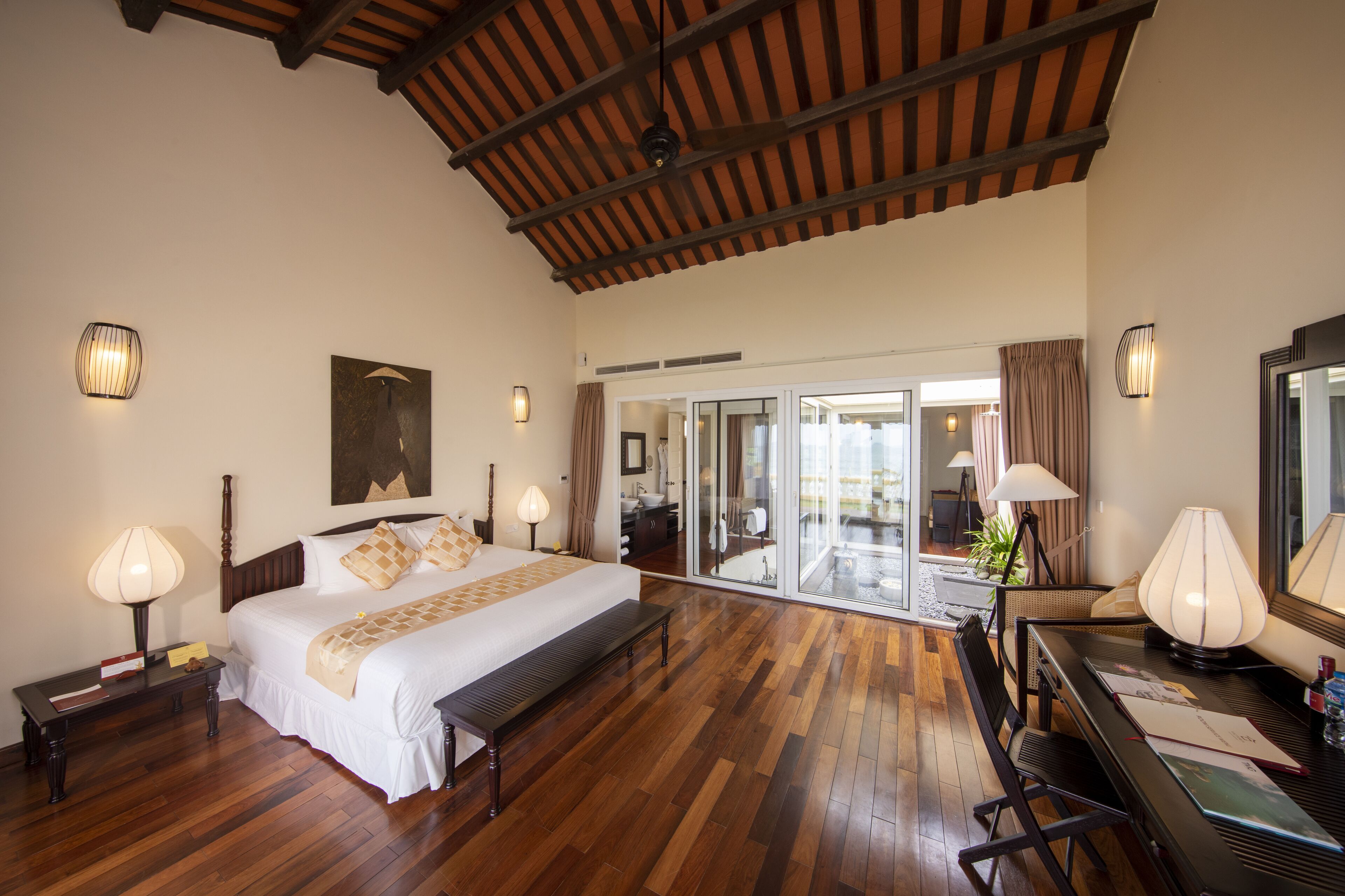 A spacious bedroom with a large bed, wooden flooring, a desk with a chair, and a view of the outside through a glass door.