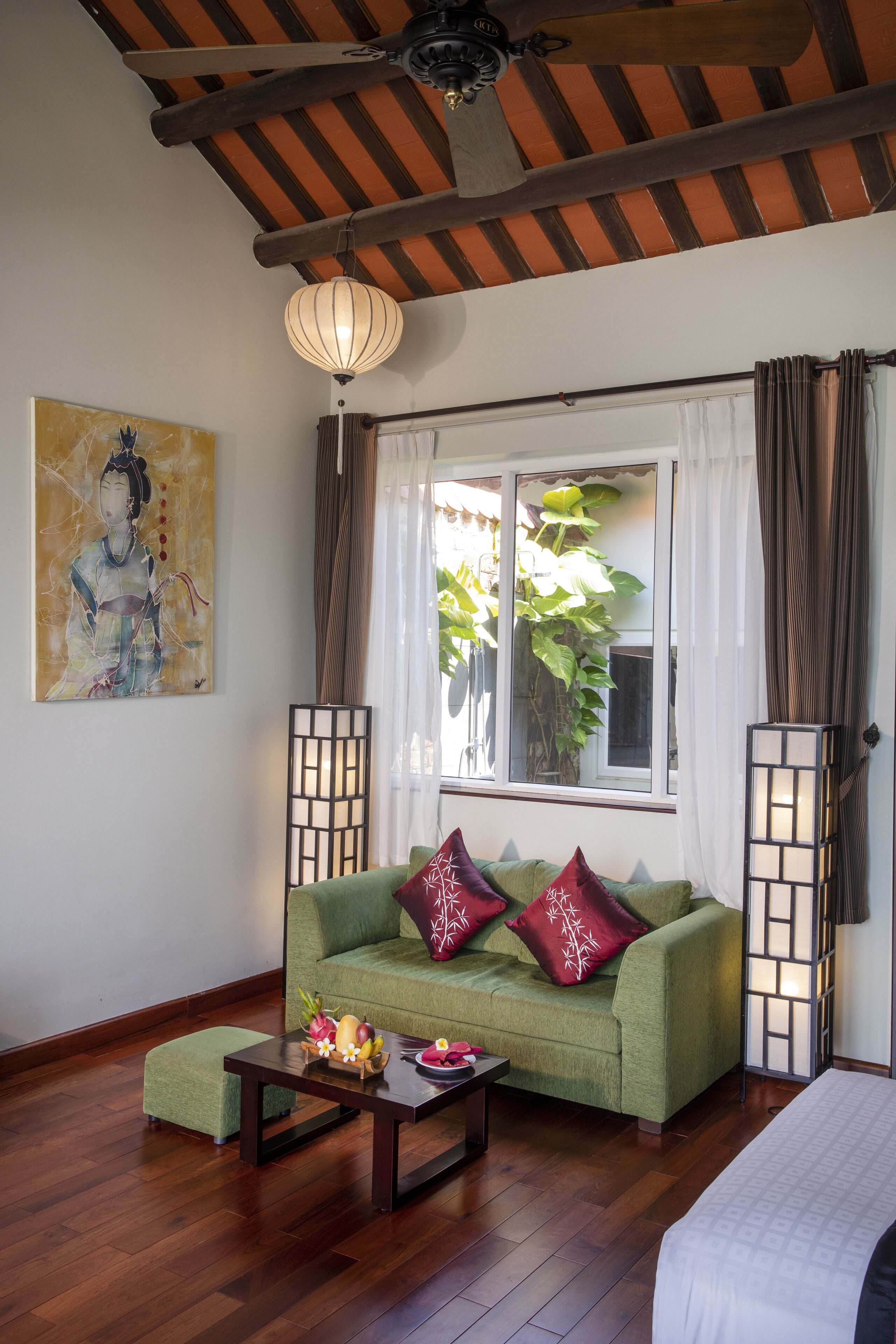 Una habitación con un sofá verde, una mesa de centro de madera, una cama, un ventilador de techo, una ventana con cortinas y un cuadro en la pared.