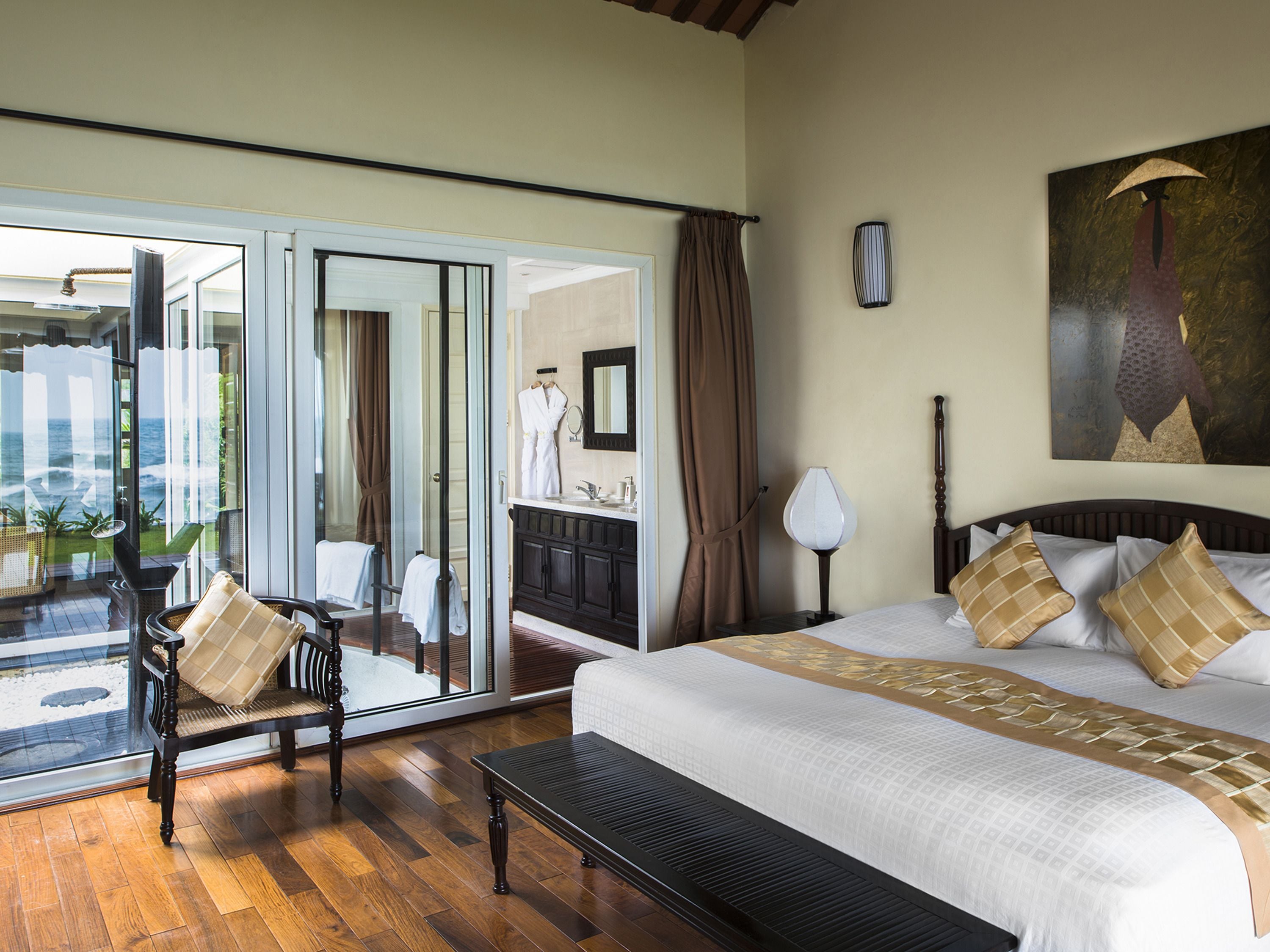 Habitación de hotel con una cama grande, un banco, un baño con espejo y vista al océano a través de una puerta de cristal.
