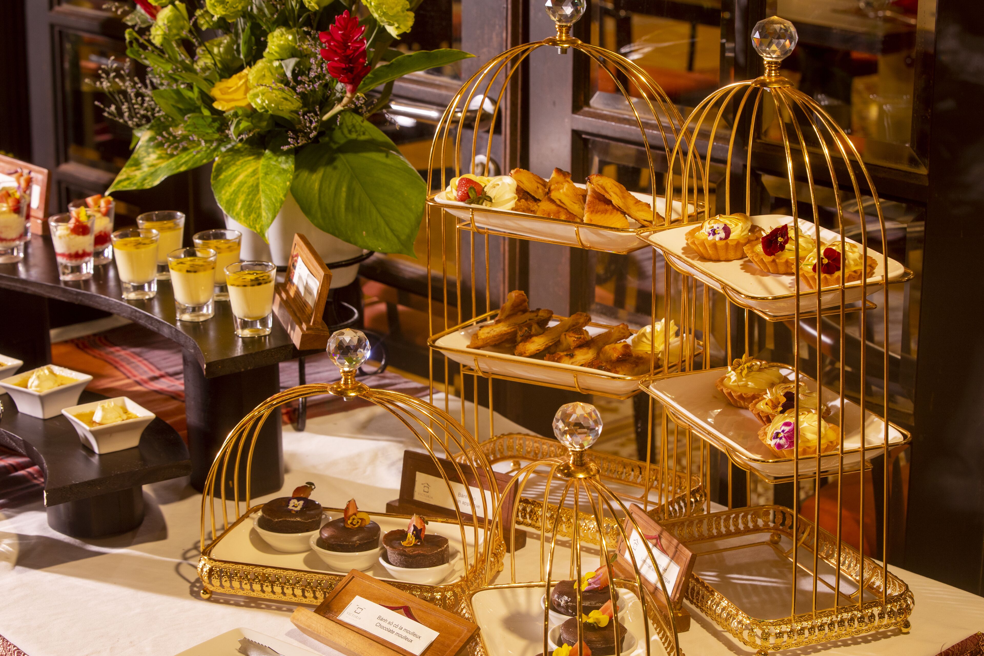 A dessert display with multiple tiers of pastries and desserts, accompanied by small glasses of a yellow beverage, set on a table with a white tablecloth.