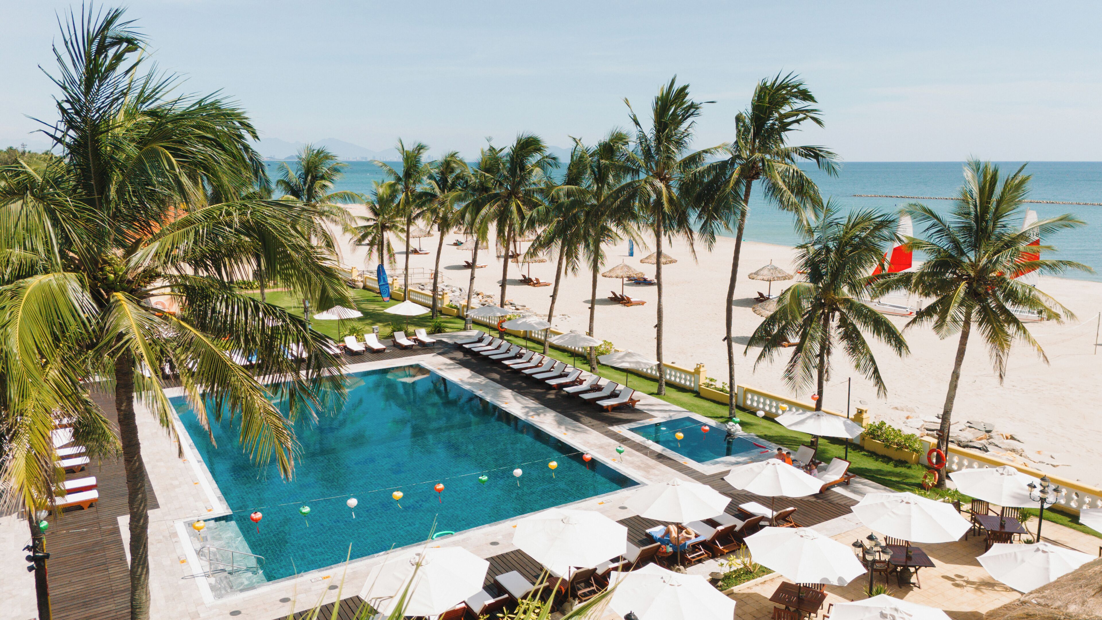 A pool area with lounge chairs and umbrellas, surrounded by palm trees and a beach.