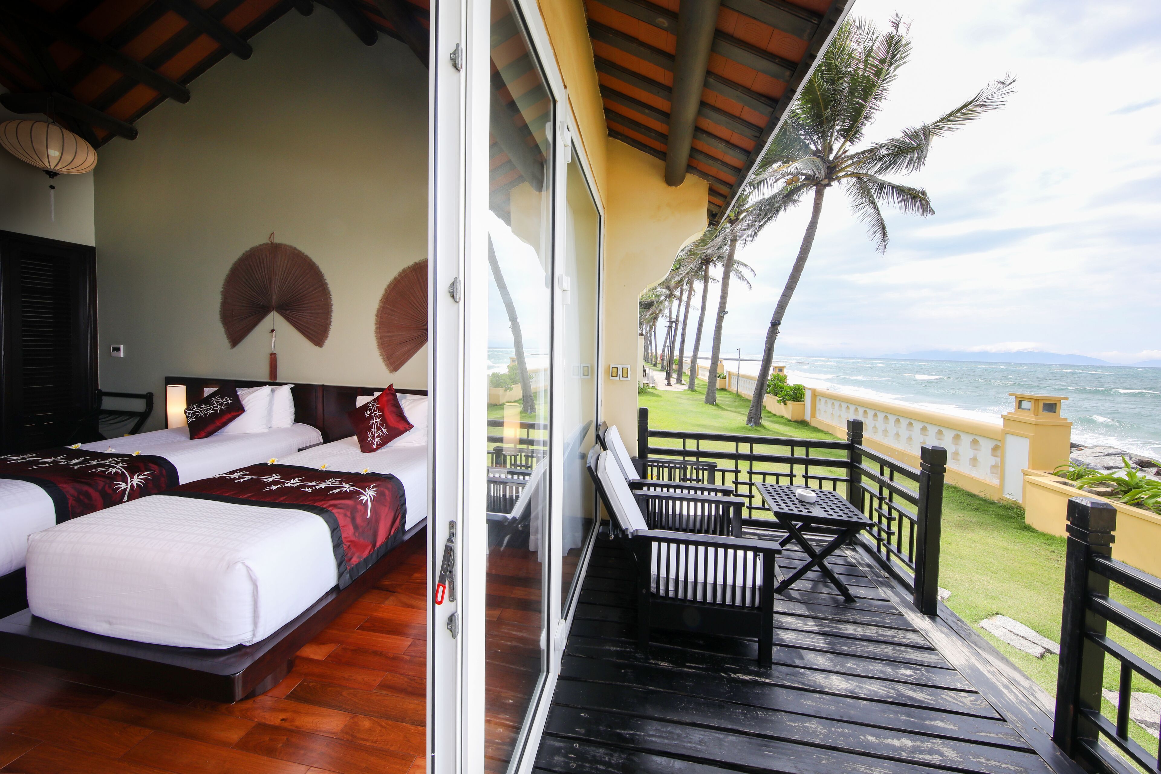 A balcony with a wooden floor and a table, overlooking a beach with palm trees and the ocean.