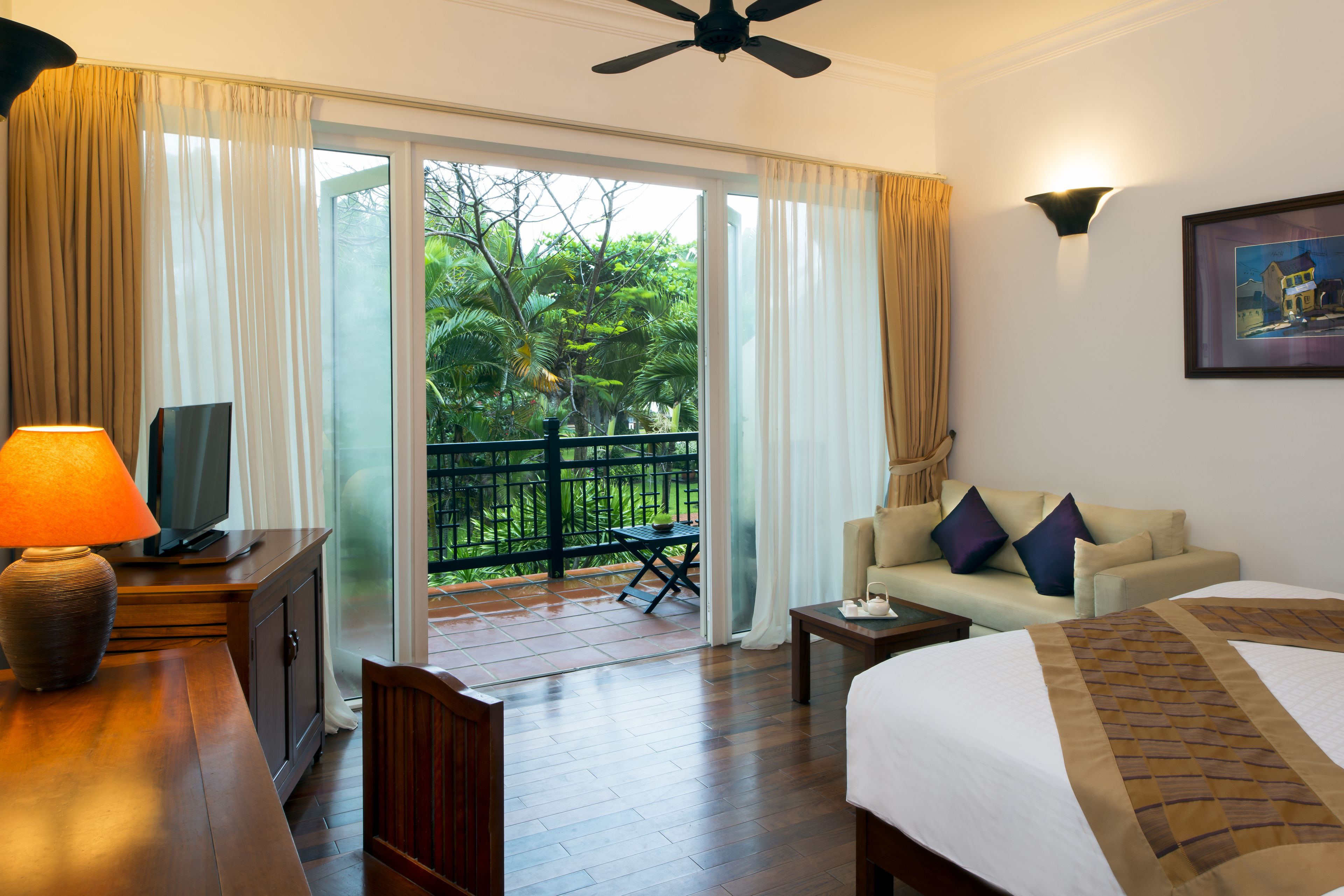 Habitación de hotel con cama, sofá, televisión sobre un mueble de madera, una mesa con lámpara y balcón con vistas a zonas verdes.