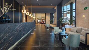 Un moderno vestíbulo de hotel con un mostrador de recepción de mármol, zona de estar con sofás y sillones, y grandes ventanales con vistas a la ciudad.